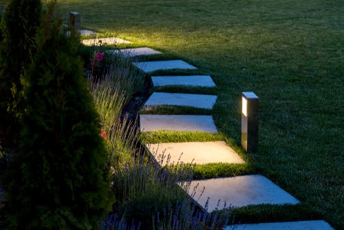 marble path of square tiles illuminated by a lantern glowing with a warm light in a backyard garden with a flower bed and a lawn copy space.